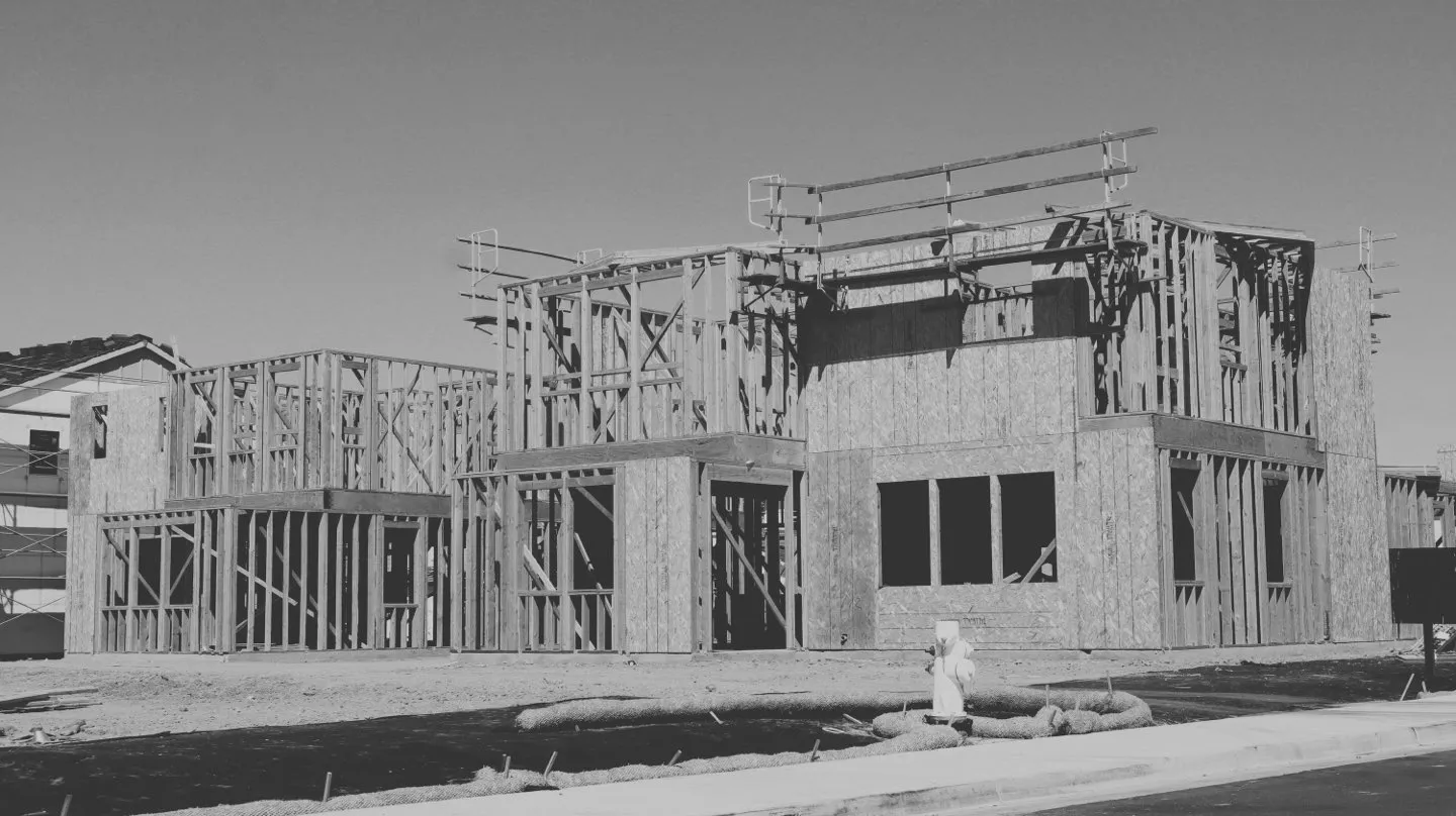 A two-storey timber building frame under construction on open land, in black and white.