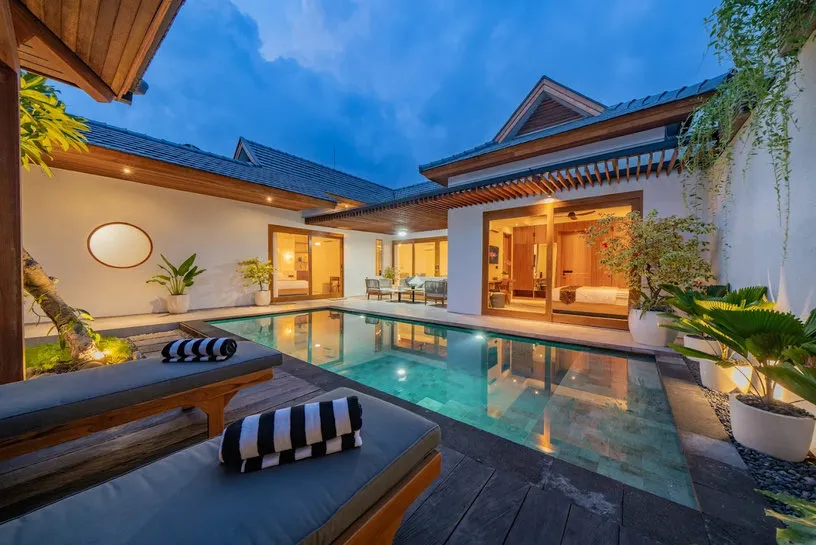 A pool and timber-roofed pavilion at dusk, with black-and-white striped cushions.