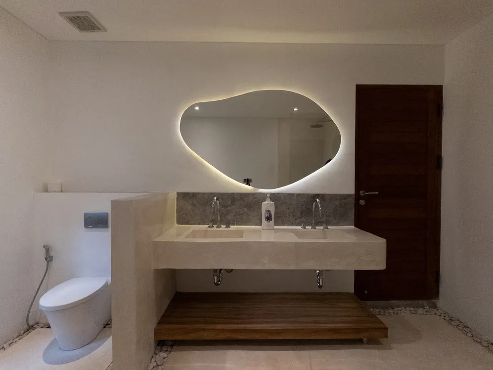 A bathroom with an illuminated organic-shaped mirror and twin basins.